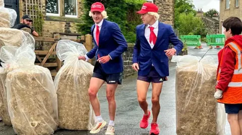 Two men dressed in blue suits and red ties sport blond wigs and red caps emblazoned with the phrase "Make America Sprint Again" in white lettering. The man on the left has his hand wrapped around a bale of hay which sits on the floor.