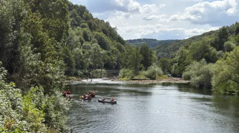 A river with at least three canoes in the bottom part of the image. Trees dominate the photo on the left and there are clouds in the sky at the top of the image.