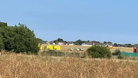 Nick Hull/BBC A field with a number of ambulances and police cars parked in it. A row of houses is in the distance.