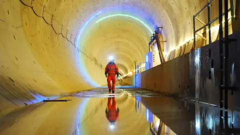 PA Media A person in high‑visibility clothing and a headlamp walks along a wet surface inside a large, illuminated rail tunnel.