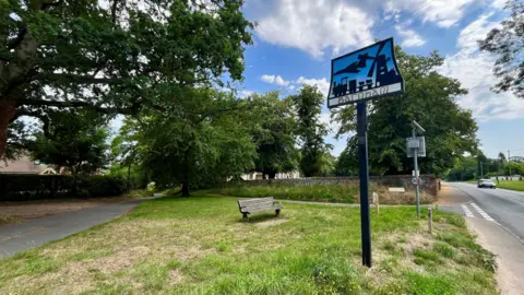 Martin Giles/BBC A village sign for Barnham is placed on a metal post on a small area of grass. There is a bench nearby.