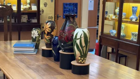 A number of painted pottery products are displayed on a table, with others in glass cabinets behind. There is also a small pile of books on the table.
