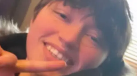 Close up picture of Demi smiling looking at the camera. She has short brown hair and wears a black coat with a fur hood. She is doing a peace pose with her one hand.