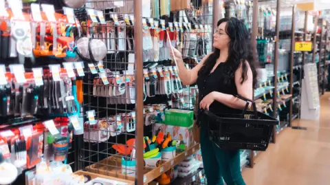 Getty Images A woman in a black tank top and black pants holds a shopping basket while shopping for cooking tools
