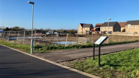 SOUTH CAMBRIDGESHIRE DISTRICT COUNCIL A plot of land partially surrounded by metal fencing in between a road and some houses. It is largely made up of mud. There are construction vehicles parked around its perimeter. 
