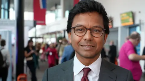 Kanishka Narayan smiles as he looks at the camera. The background is blurred but he appears to be at a conference or event.