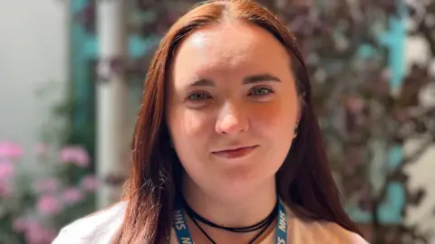 Dr Viktoriya Betsal. She has long dark red hair and is pictured looking directly at the camera and smiling. She has a blue NHS lanyard around her neck.