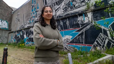 Ainhoa Burgos Aguilera, a woman in a green jumper, stands arms-crossed in front of a grey wall covered in graffiti.