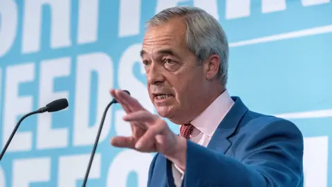 Nigel Farage looks up and away from camera with two small microphones in shot against a light blue background, as he delivers a speech at a press conference on 10 July in London.