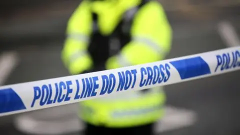 BBC Generic image of a police tape cordon, with an office in high-vis, out of focus in the background