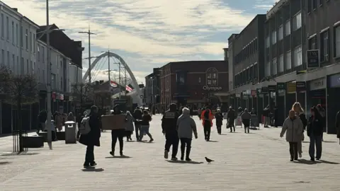 People walk along a pedestrainised street in Bolton town centre, lined with shops. A modern white arch features in the background.