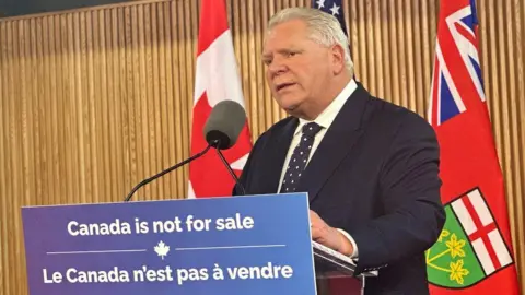 Doug Ford in a black suit stands behind a podium that reads "Canada is not for sale". Behind him is both the blue stars of a US flag, Canadian flag, with its red maple lead, and the red Ontario flag that also features the Union Jack.