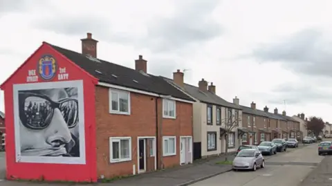 A screengrab of a Google Streetview image of Templemore Street in east Belfast. It shows a row of terraced houses with different brick exteriors and several cars lining the pavement. There is a large black and white loyalist mural on the gable wall of the first house. It shows a person wearing large black sunglasses with a cloth or fabric covering their mouth. Above the image there is a UDA crest and writing which says "Dee St, 2nd Batt."
