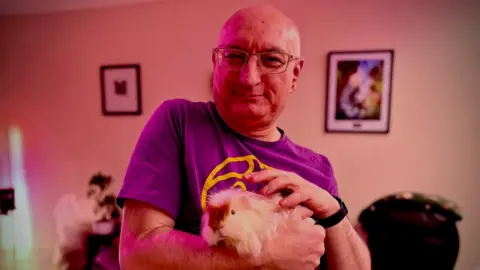 A bald man wearing glasses holds his pet guinea pig, who's mostly white with a brown splodge on its face, in his arms. 