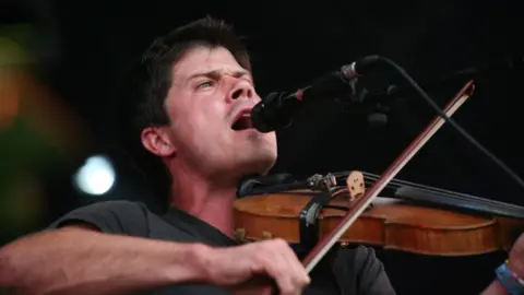 Seth Lakeman sings or shouts into a microphone, while holding a violin against his chin. He has dark hair and a dark T-shirt. The image is taken from below looking up at him.