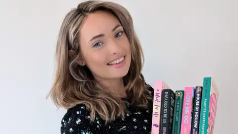 A woman holding books while smiling at the camera. She has a black top with a floral pattern on it.