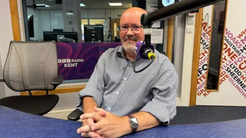 A man sat in a radio studio with a radio mic partially covering his face. He is wearing a blue and white shirt and a pair of glasses. 