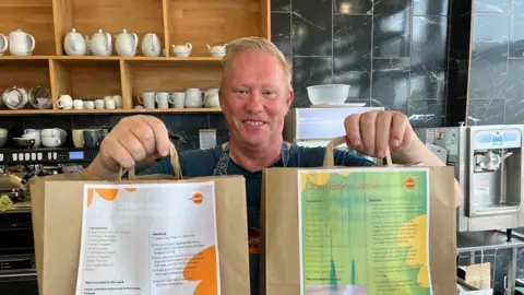 A man, Barry Ashley, stood in a cafe holding two paper bags out in front of him.