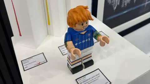 Contributed A view of the Ed Sheeran Lego figure from above. The whole figure can be seen. It is sitting on a white display table with an information card in front of it detailing information about the artist and creation. 