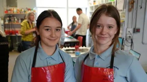 Qays Najm/BBC Amelie and Tabitha. Two schoolgirls in their pale blue school uniform wearing red aprons. They are both looking directly at the camera and smiling.