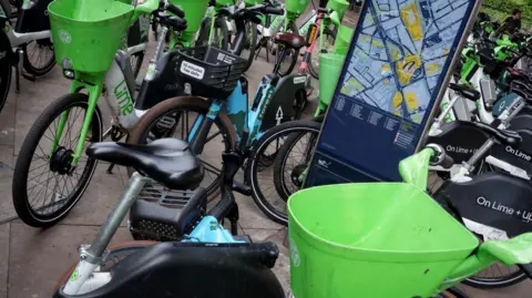 Lots of blue and green e-bikes parked up and a screen showing Hammersmith on a map 
