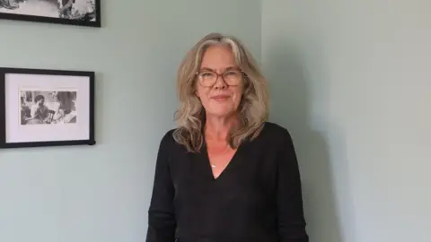 Society of St. James Tania is white and is wearing brown-framed glasses. She has fair hair and is wearing a black top with a silver necklace. She is standing in front of a wall and to her right is a framed black and white photograph of two women.