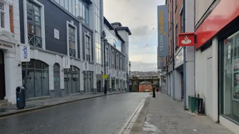 Image shows a wide shot of Butcher Street. A blue and white building with signs for apex is visible on the left and a red building beside a grey building signed maldron Hotel can be seen on the right side. The street is empty and a cloudy sky can be seen in the background.