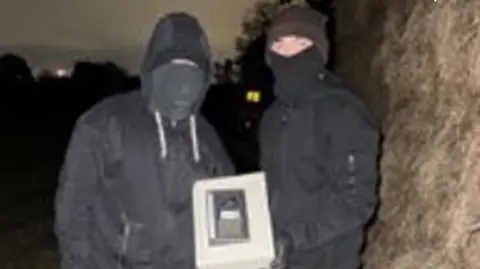 Cheshire Police Two of the gang members wearing dark clothes and face coverings and with their hoods up hiding their faces hold up a safe. They are outside in the county in the dark.