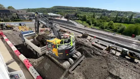 Building works taking place on the railway line near Huddersfield station. There is a digger in the foreground and a railway rack in the background, with moors in the background as well. 