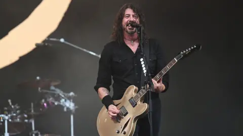 Dave Grohl and Foo Fighters performing on the Pyramid Stage at Glastonbury Festival 2023. He is smiling holding a guitar and has drums in background