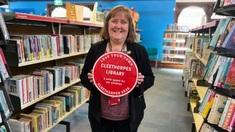 A woman smiles, with shoulder-length brunette hair, holding the red plaque which reads: 'Love your town, Cleethorpes Library, A safe space for children, Cleethorpes 2026'.
She is photographed down an aisle of books in a library.