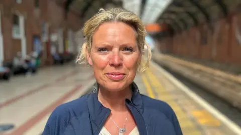 Katie Swinburne looking into the camera with a slight smile. She has blonde hair which is tied back and is wearing a blue jacket, silver necklace and white top underneath. She is standing in a train station on a platform which is out of focus.