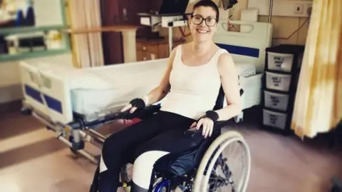 Stevie O'Brien A woman with short dark hair and glasses sits smiling in a wheelchair in a hospital ward.
