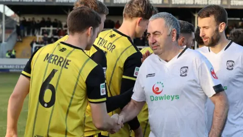 Bulls News A man with grey-white hair wearing a Hereford FC kit moves along a line of AFC Fylde players, shaking hands with them.