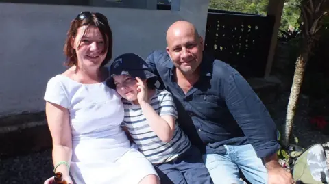 Nicky Wake A family of three sit in a garden in sunny weather.