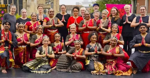Graham Day A large group picture of the dance company on a stage outdoors following a public performance. All dancers are wearing Bharatanatyam costumes and have their finger tips painted red so the audience can better see their precise hand movements.