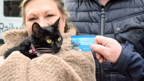 South Western Railway A black cat with yellow eyes staring at her blue railcard. She is being held by a woman in a beige coat 