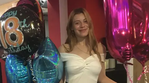 A young woman with blonde hair and wearing a white dress smiles as she holds a number of balloons, one of which says "happy 18th birthday". 