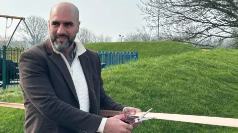 Gavin Kermack / BBC A bald man with a salt-and-pepper beard, wearing a brown jacket over a white cardigan, holds a pair of scissors against a ribbon in a park.