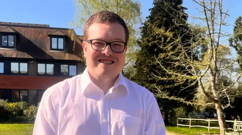 Jack wears a light pink shirt and dark trousers. He has closely cropped brown hair and black rimmed glasses. He smiles and his hands are clasped together. He is standing in a green area outside a building.