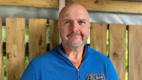 Andy Jones, smiling at the camera. He has a bald head and a short grey beard. He is wearing a blue sweat top.