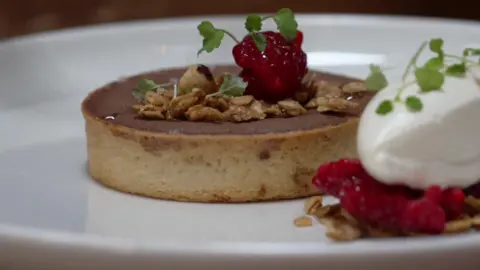 A close up of a molten chocolate tart with an oat crumble served on a white plate. It has a raspberry on top of it.