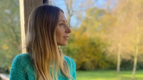 Ali Harris Ms Harris with long, straight hair, sitting outdoors. She is wearing a turquoise jumper and is turned slightly to her right, so her face is in profile. Behind her is a backdrop of trees with golden and green leaves. The lighting is soft and natural.