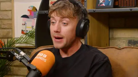 A man, Roman Kemp, wears a black top and sits on a sofa in front of a Sounds branded microphone