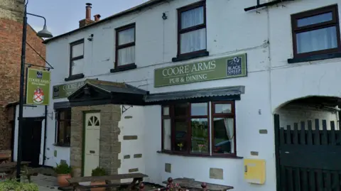 A traditional white‑painted pub called the Coore Arms, with dark brown window frames, a small stone porch at the entrance, and hanging signs for “Coore Arms Pub & Dining.” Wooden benches and potted plants sit outside on a quiet village street.