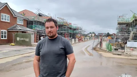 Nathan Long stand on the street in front of a houses with scaffolding and builder's fencing. 