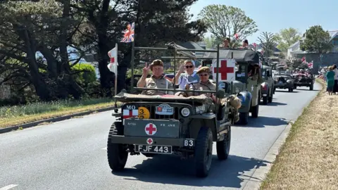 A picture of people posing in a vehicle that was parading in the cavalcade. The vehicles are green in colour on a grey road.