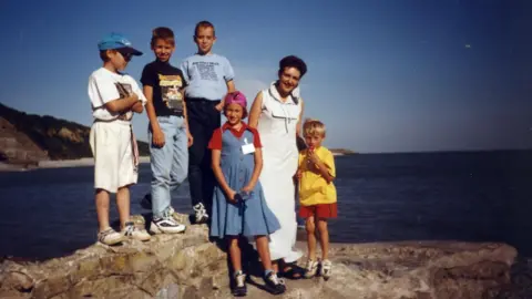 Anton Honchanka Anton central in a blue t shirt. He is flanked by three boys, a girl and a woman. They are stood on a rock with the sea and coast behind them.