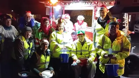 Devizes Lions 11 people stood in an outdoor setting with a man dressed up as Santa. Some are in high-vis jackets, others wear santa hats and several hold charity collection buckets.