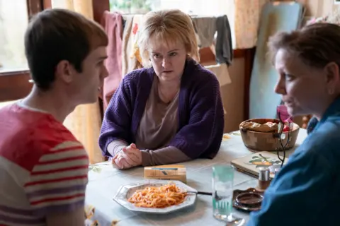 BBC/Mam Tor Productions/Anne Binckebanck A teenager wearing a red and white top sits at a kitchen table. A blonde woman wearing purple and a brunette woman wearing blue speak to him.